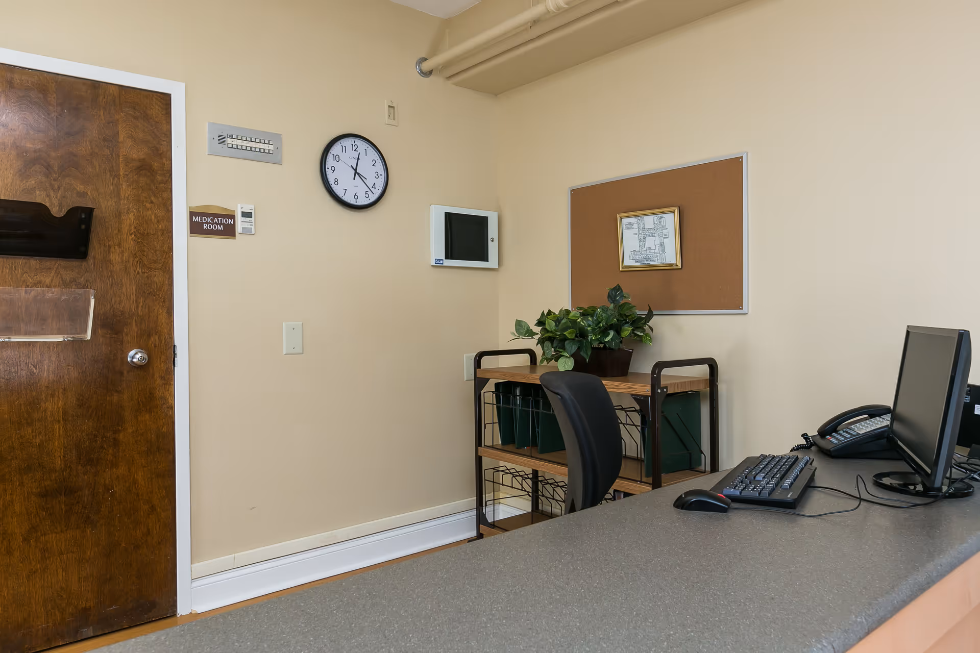 Interior view of a medication room in a nursing facility with a wooden door labeled 'Medication Room', a wall clock, a computer workstation with a keyboard, mouse, and telephone, a chair, a small shelf with green bins, a potted plant, and a cork bulletin board with a framed paper pinned on it.