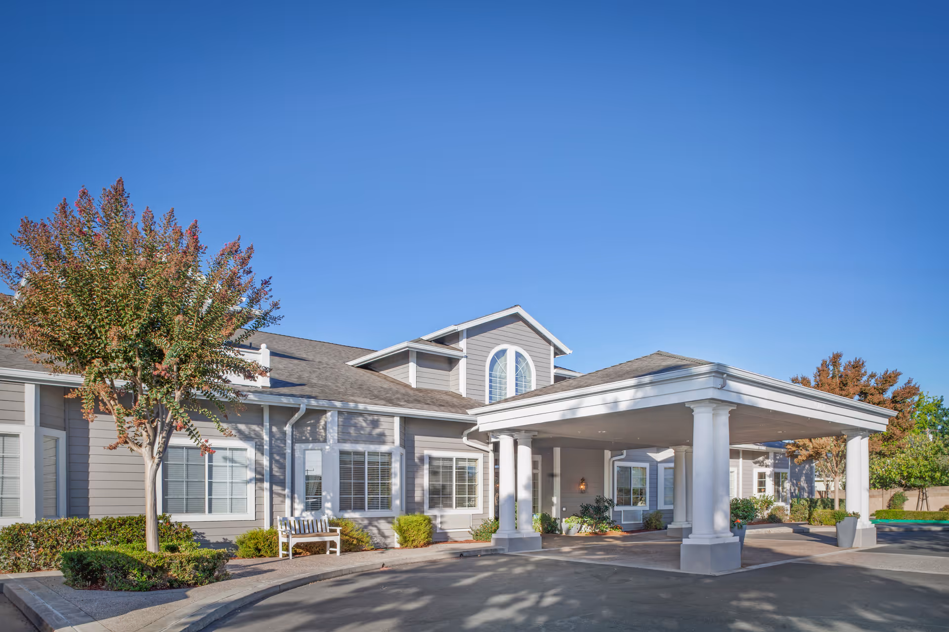 Exterior view of a senior living facility building with a covered entrance supported by white columns. The building is light gray with white trim and has multiple windows. There are trees and shrubs around the entrance area under a clear blue sky.