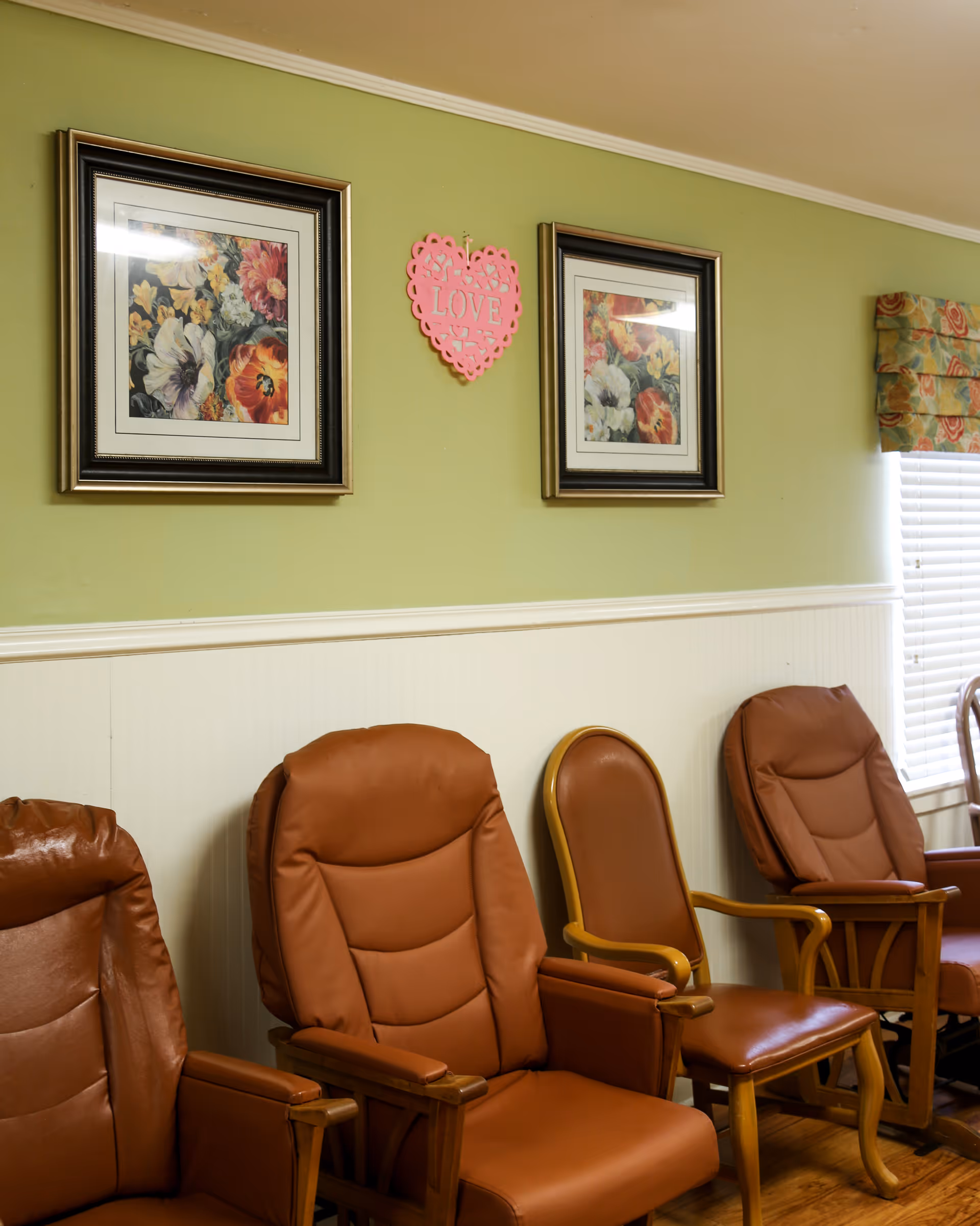 A row of brown cushioned chairs with wooden armrests lined up against a light green wall with white wainscoting. Above the chairs, there are two framed floral paintings and a pink heart-shaped decoration with the word 'LOVE' on it. A window with floral-patterned curtains is partially visible on the right side.