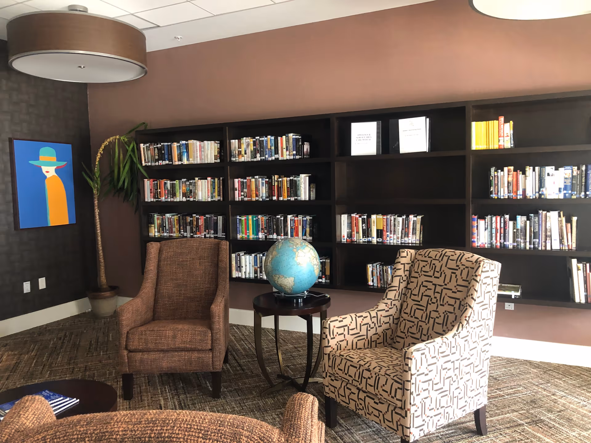 A cozy reading area in a senior living community featuring two upholstered armchairs, a small round table with a globe on it, and a large dark wood bookshelf filled with books. The walls are painted in warm tones, and there is a colorful modern painting and a potted plant in the corner.