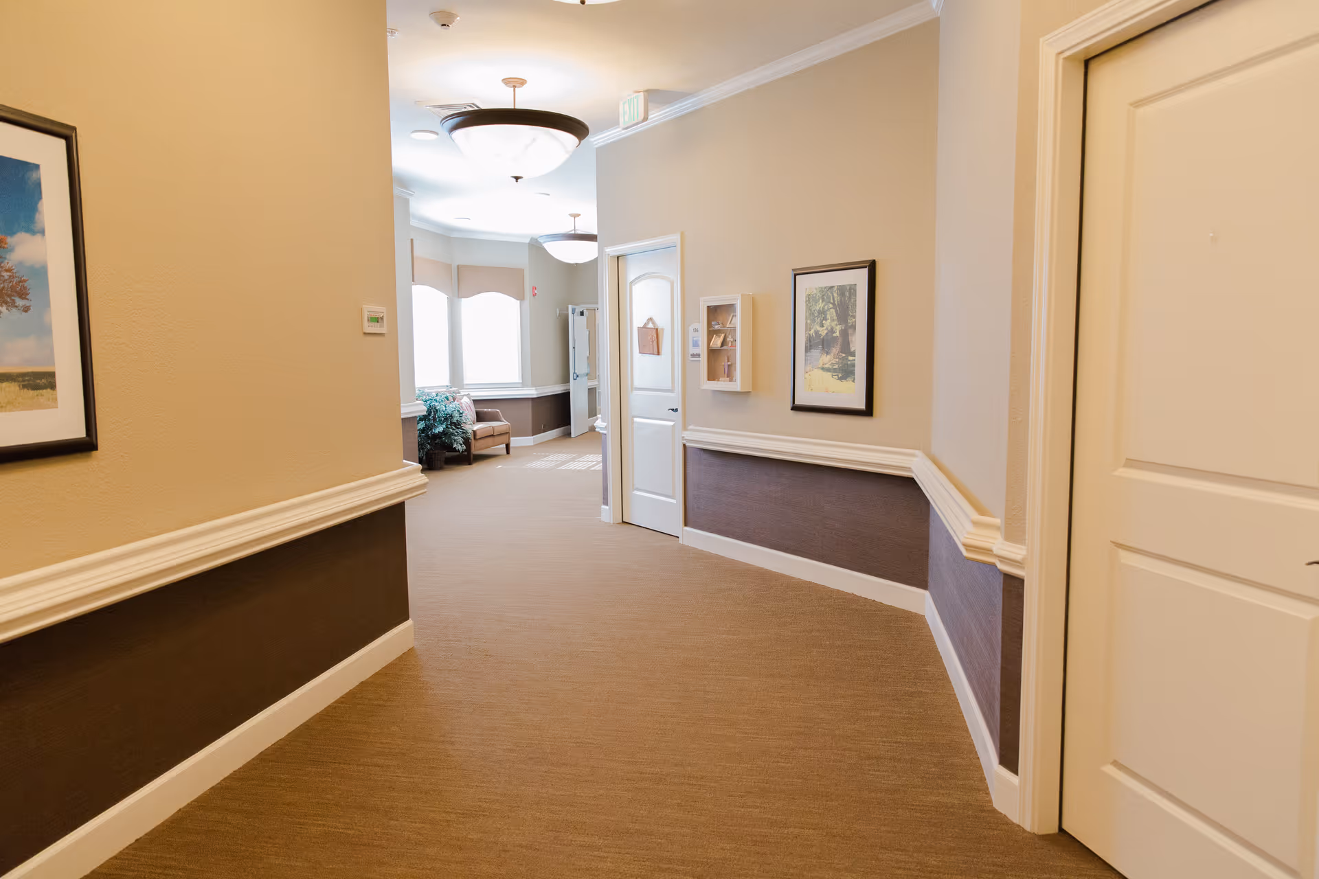 Well-lit carpeted hallway in a senior living facility with framed artwork, white doors, and seating visible at the far end.