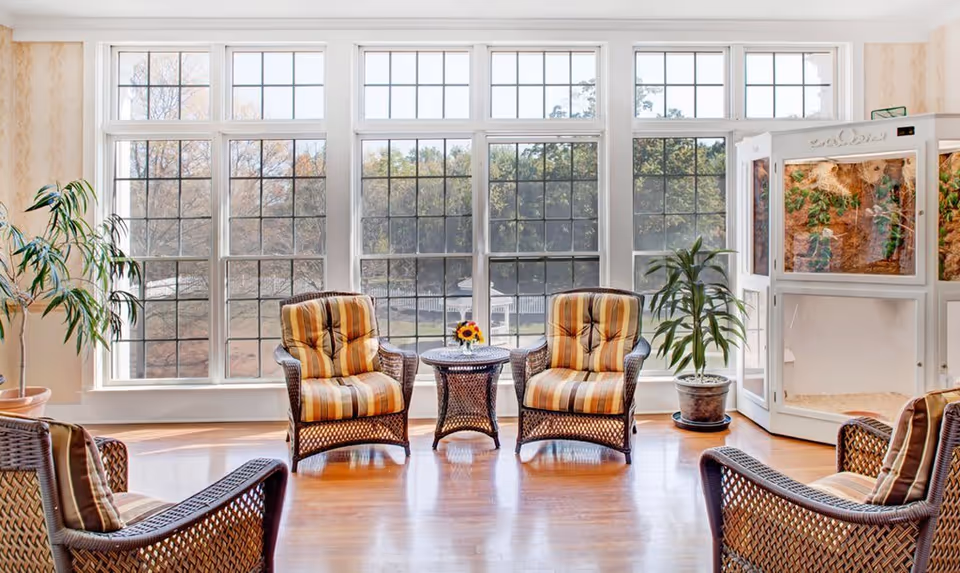 A bright and airy sitting area with large windows letting in natural light. The room features four wicker chairs with striped cushions arranged around a small wicker table with a vase of flowers. There are two potted plants near the windows and a large white enclosure on the right side of the room.