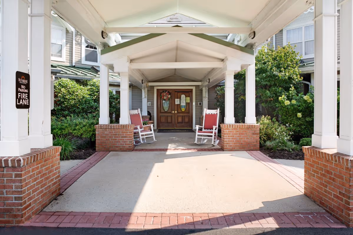Covered entrance to a building with a peaked roof supported by white columns on brick bases. Two white rocking chairs with red cushions sit on either side of the entrance door, which is wooden with glass panels and decorated with a wreath. Green shrubs and plants surround the entrance area. A sign on the left column reads 'Sunrise No Parking Fire Lane.'