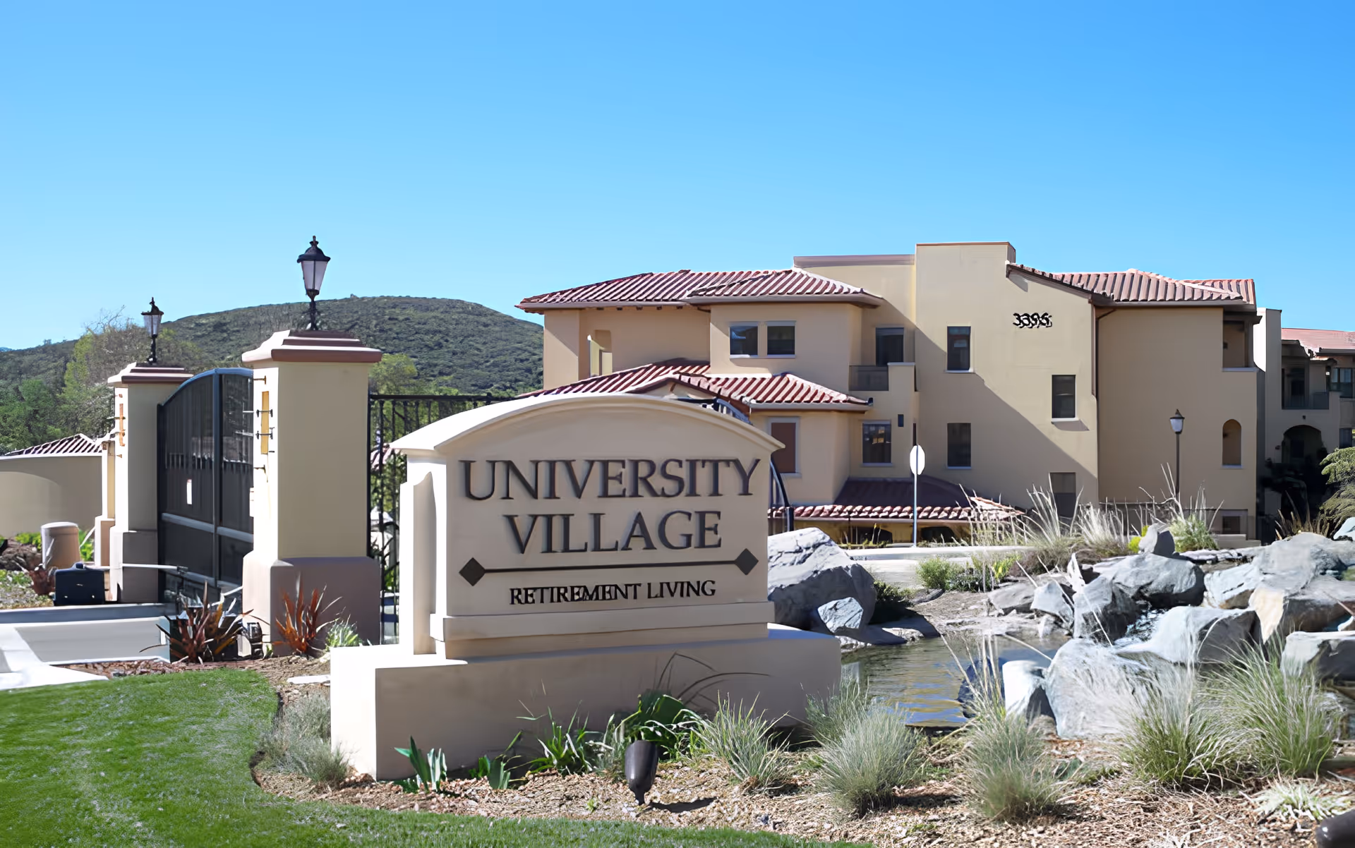 Stone sign reading 'University Village Retirement Living' at the landscaped entrance to a Mediterranean-style retirement community with buildings, rocks, and a pond.