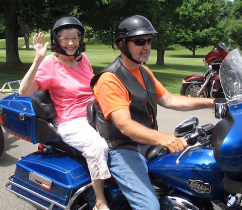 An elderly woman wearing a pink shirt and white pants is sitting on the back of a blue Harley Davidson motorcycle, waving and smiling. A man wearing an orange shirt, black leather vest, sunglasses, and a helmet is driving the motorcycle. They are outdoors with green grass and trees in the background.