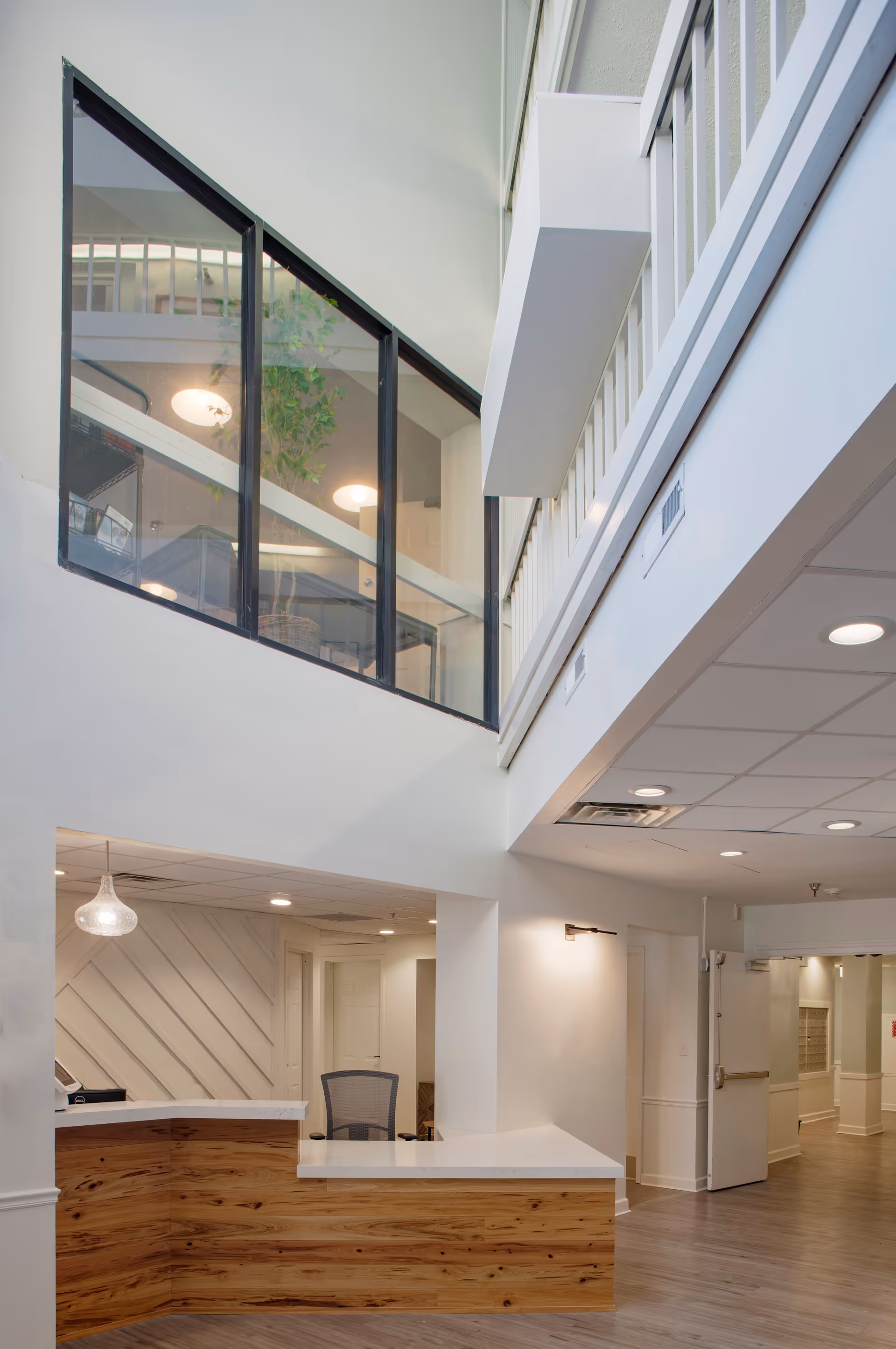 Interior view of a senior living facility reception area with a wooden front desk and white countertop. The space features high ceilings with a second-floor balcony enclosed by glass panels. The walls and ceiling are painted white, and the floor is a light wood tone. There is a hallway leading to other rooms and a modern light fixture hanging above the reception desk.