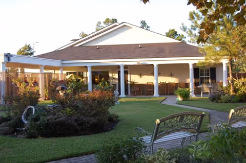 Outdoor view of a single-story building with a covered porch area featuring several chairs and tables. The foreground includes a well-maintained lawn, garden beds with shrubs, and decorative benches along a paved pathway. Trees and clear sky are visible in the background.