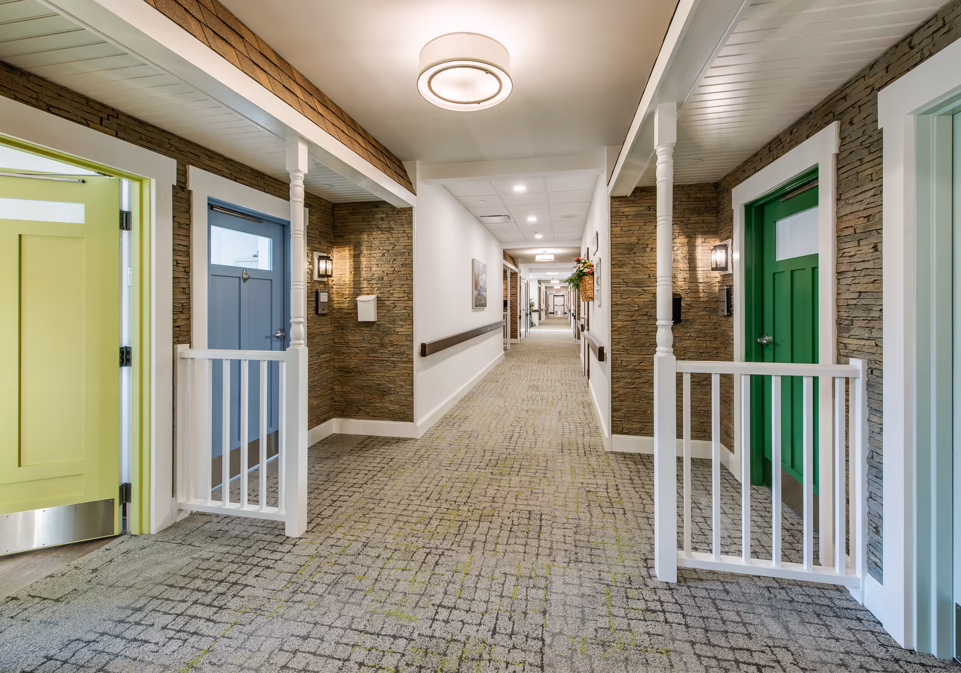 A long, carpeted interior hallway with blue, green, and yellow doors flanked by porch-style railings and stone-accent walls.