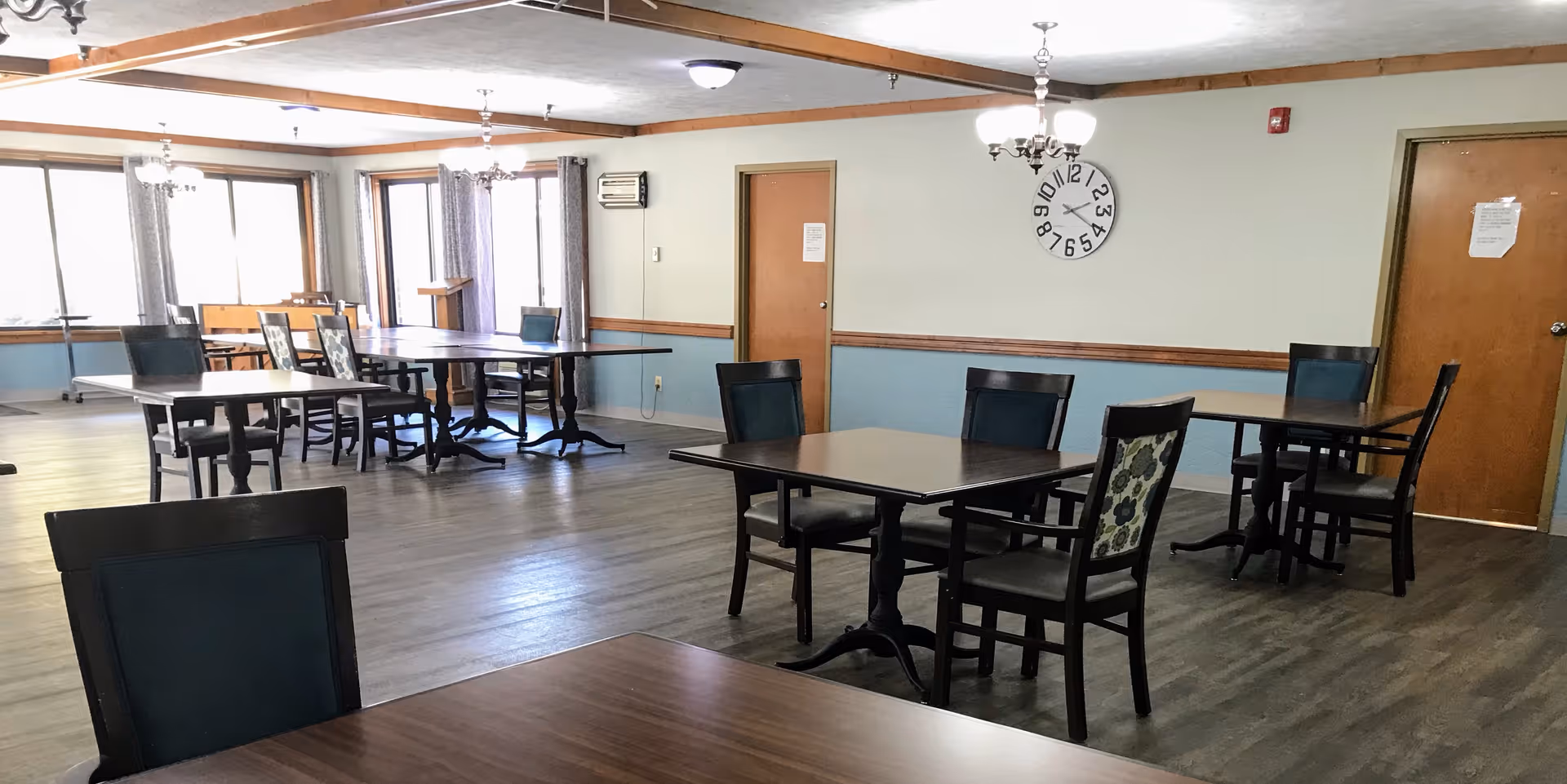 A spacious dining room with multiple dark wood tables and chairs arranged neatly. The room has large windows with curtains allowing natural light to enter. There is a wall clock and two closed wooden doors on the far wall. The floor is covered with wood-like vinyl flooring and the ceiling has wooden beams and light fixtures.