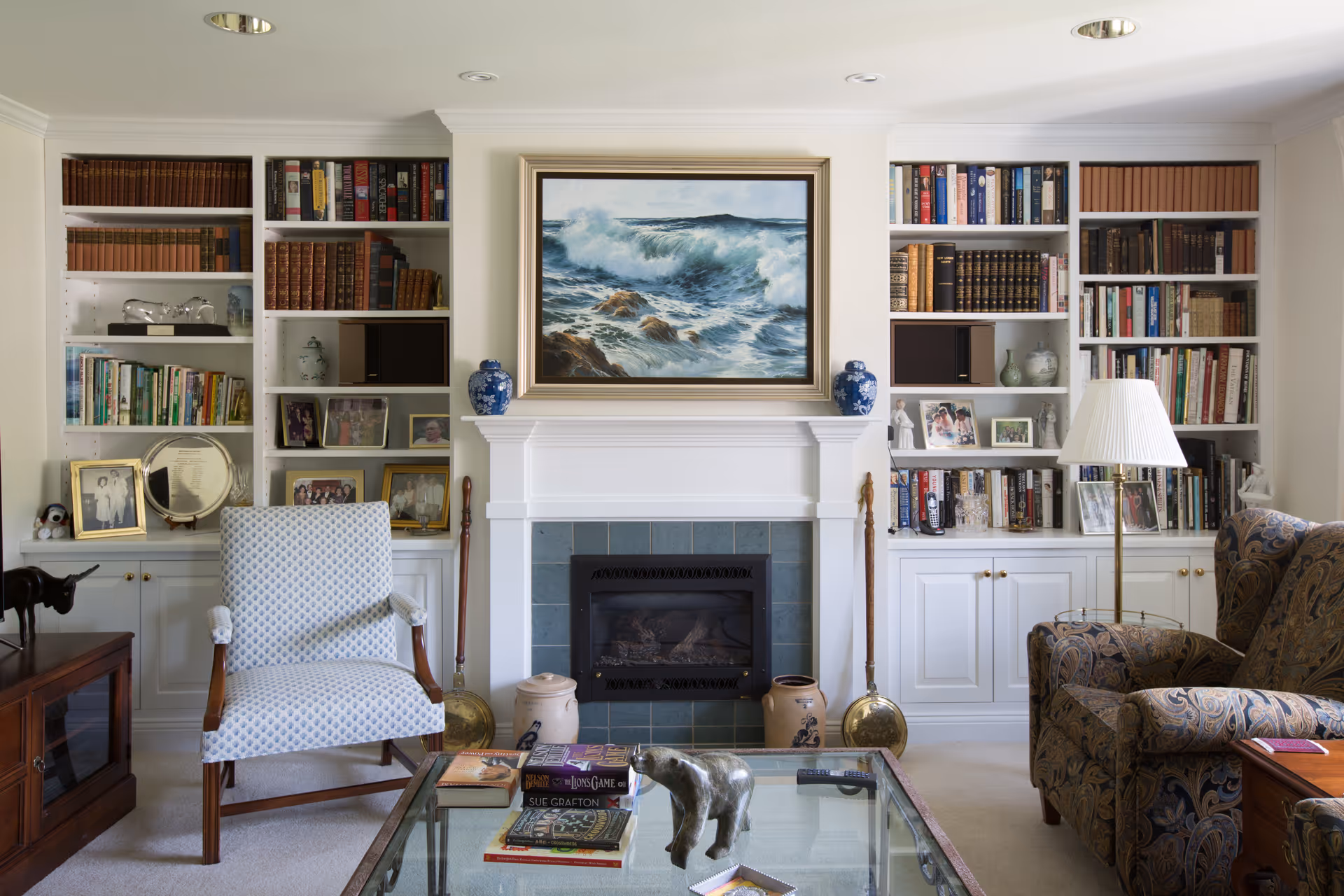 A cozy living room with a white fireplace in the center, flanked by built-in bookshelves filled with books, framed photos, and decorative items. Above the fireplace is a framed painting of ocean waves. In front of the fireplace is a glass coffee table with books and a small bear figurine. There is a patterned armchair on the right with a floor lamp beside it, and a light blue patterned armchair on the left. The room has a warm and inviting atmosphere.
