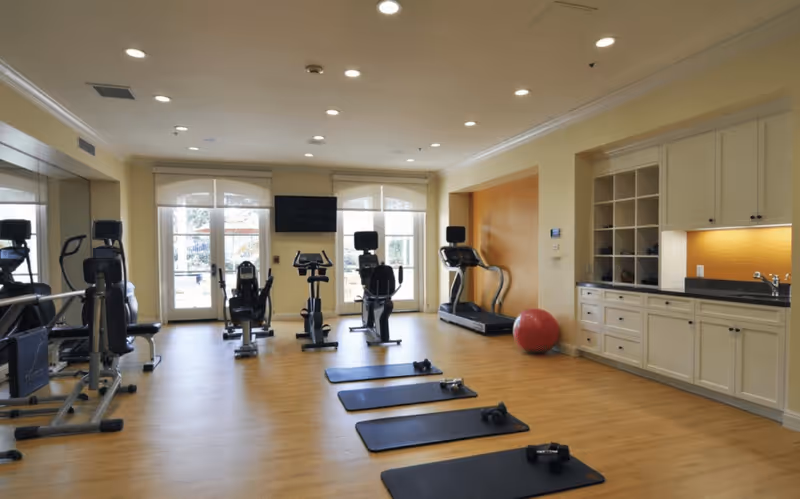 Bright fitness room with cardio machines, floor mats and dumbbells, a stability ball, and built-in cabinets.