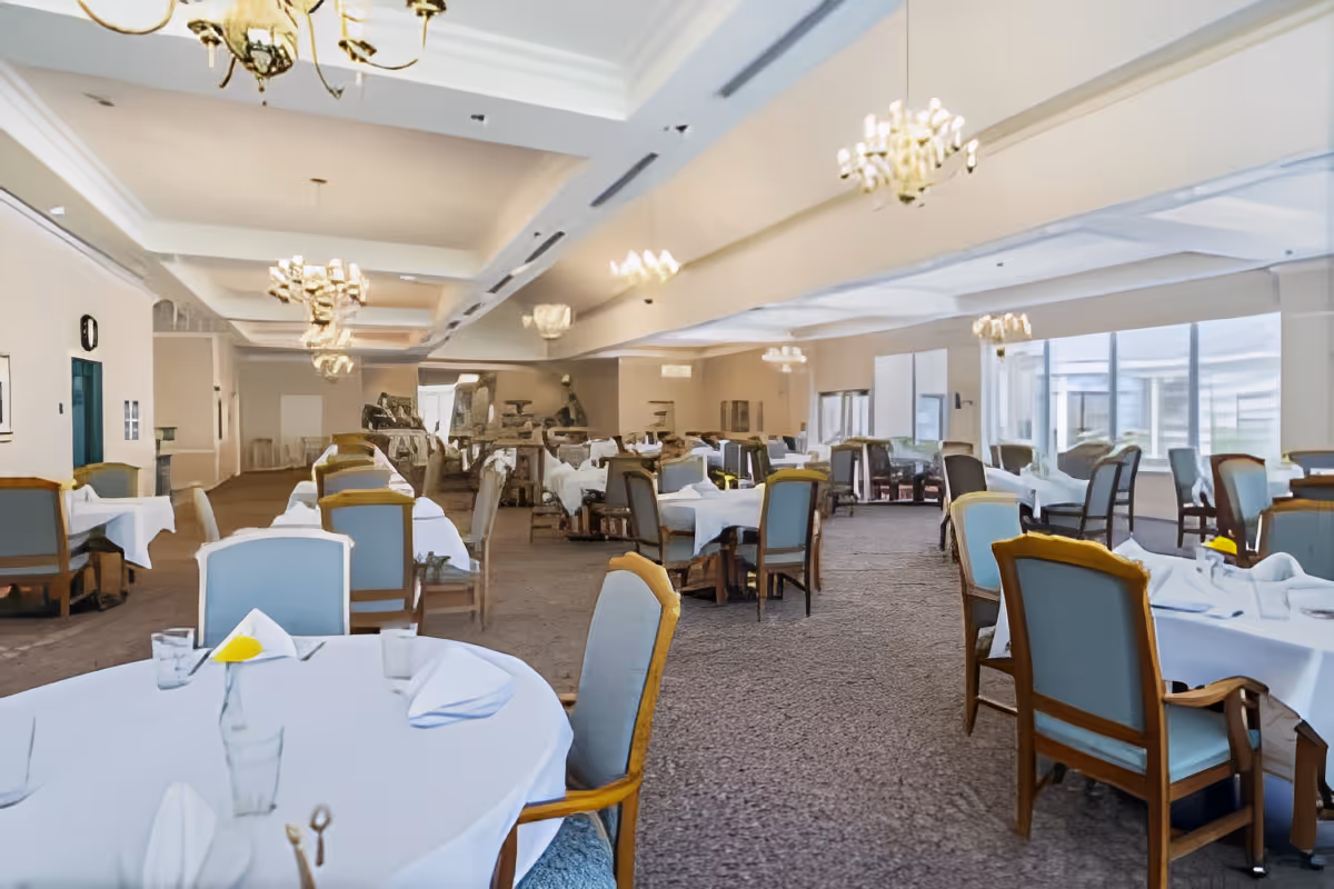 A spacious dining room with multiple round and rectangular tables covered with white tablecloths, set with glasses, napkins, and silverware. The room features upholstered chairs with wooden frames, large windows letting in natural light, and several chandeliers hanging from the ceiling.