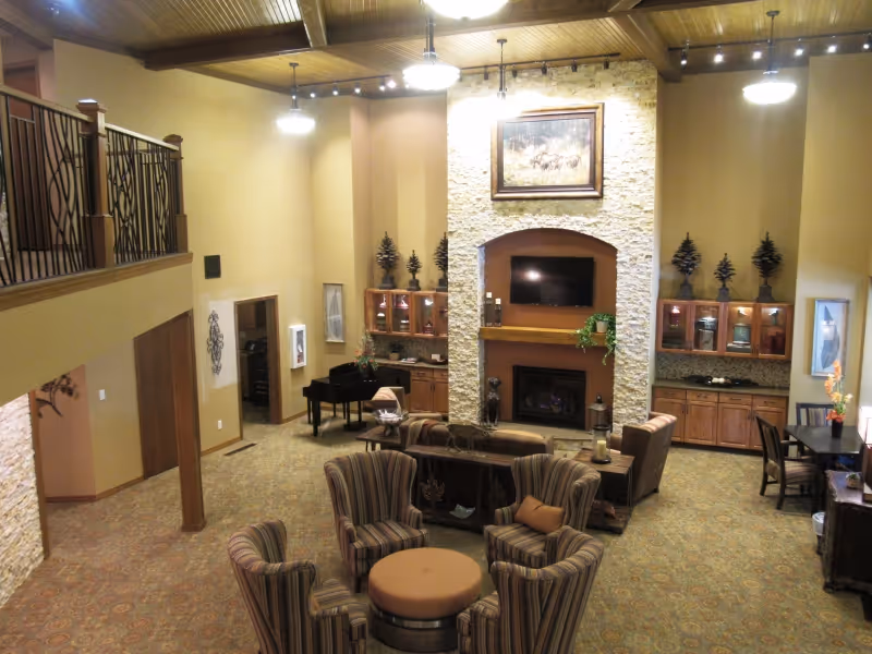 Spacious living room area in Creekside at Elfindale Independent Living with high wooden ceilings, a stone fireplace with a mounted TV above it, several armchairs and sofas arranged around a central ottoman, a grand piano in the corner, and wooden cabinets with decorative items. The room is warmly lit with hanging lights and has a carpeted floor.