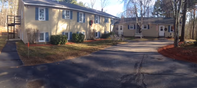 Exterior view of a two-story beige residential building with multiple windows, surrounded by trees and a paved driveway. There is a small patio area with chairs near the entrance, and the scene is set in a wooded area with clear skies.