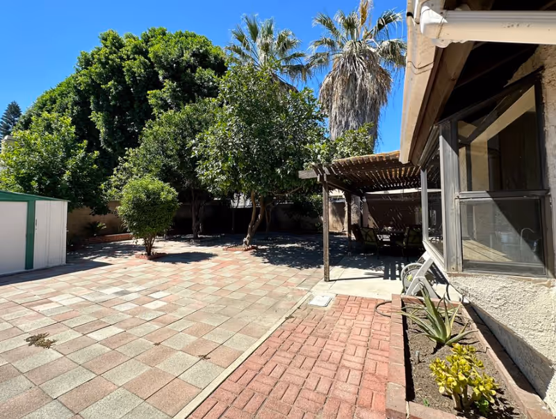 Outdoor patio area with tiled flooring and a small garden bed with plants next to a building. There is a pergola with outdoor seating underneath, surrounded by various trees including a palm tree, under a clear blue sky.