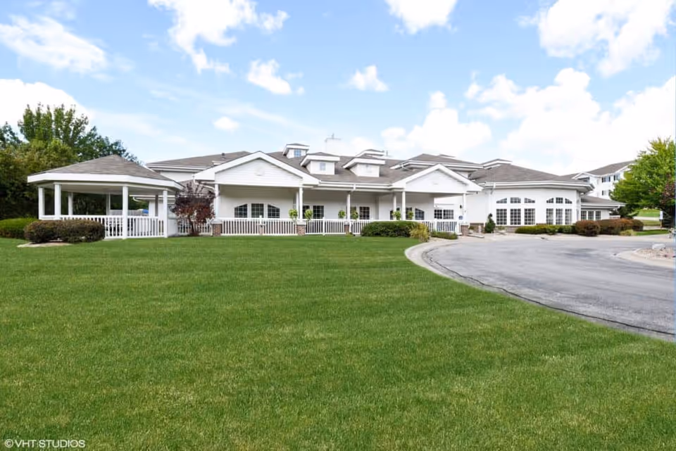 White single-story senior living facility building with a covered porch and gazebo, a large green lawn and curved driveway under a blue sky.
