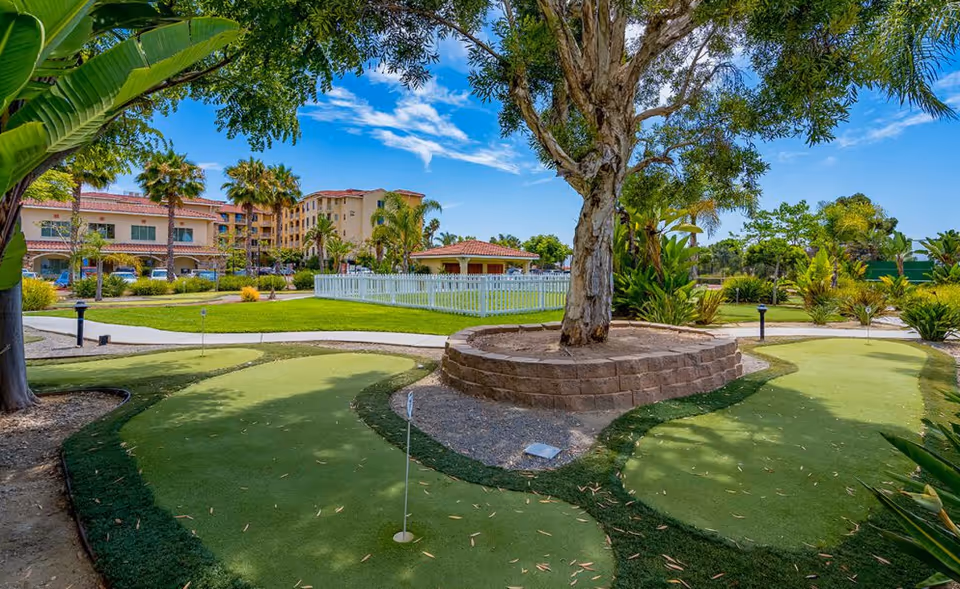 Outdoor putting green with a central tree in a raised stone planter, surrounded by landscaping and a multi-story senior living building in the background.
