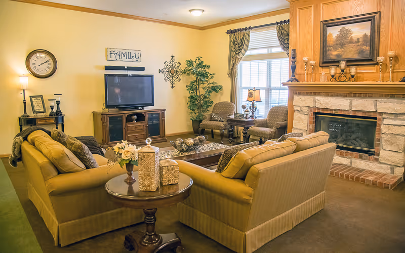 A cozy living room with two beige sofas facing a wooden TV stand with a flat-screen TV. The room features a stone fireplace with a painting above it, a round wooden side table with decorative items, two armchairs near a window with curtains, a floor lamp, and wall decorations including a clock and a 'FAMILY' sign.