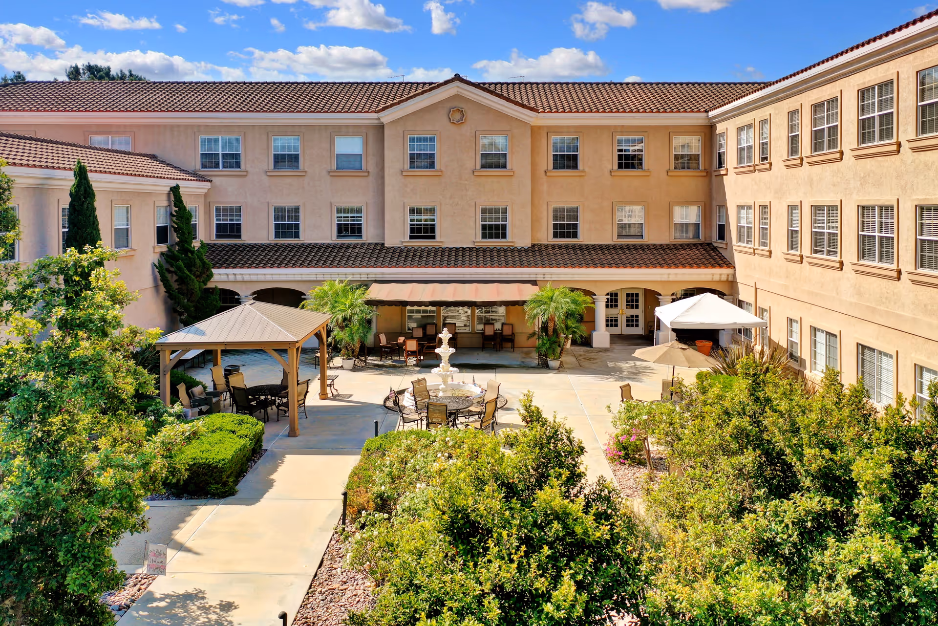 Outdoor courtyard area of a senior living facility with a central fountain, surrounded by patio tables and chairs, shaded seating areas under gazebos, and lush greenery. The courtyard is enclosed by a three-story beige building with multiple windows under a clear blue sky.