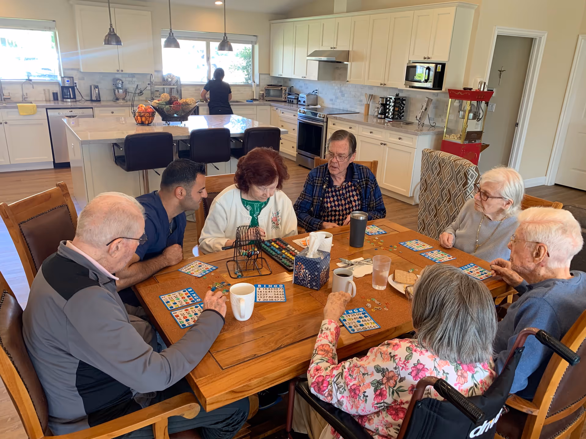 Several elderly residents sit around a wooden table in a kitchen/dining area playing bingo.