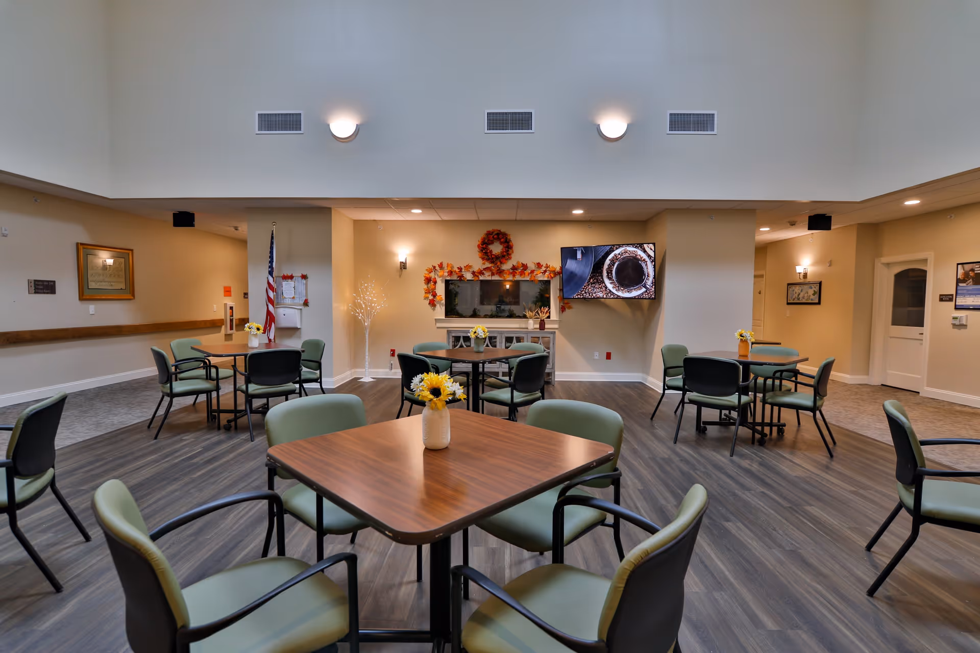 A spacious senior living common area with multiple square wooden tables surrounded by green cushioned chairs. The room has wood flooring, neutral-colored walls, and soft lighting. A flat-screen TV is mounted on the wall above a decorative fireplace adorned with autumn-themed wreaths and garlands. An American flag stands in the corner, and small flower vases decorate the tables.