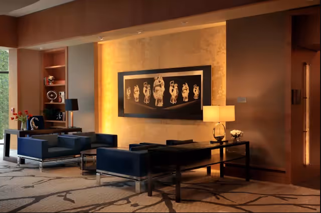 A modern living room area with dark blue armchairs and a black coffee table. The room features a large framed artwork of classical sculptures on a warmly lit wall. There is a table lamp on a side table and built-in shelves with decorative items. The floor has a patterned carpet and a large window letting in natural light.