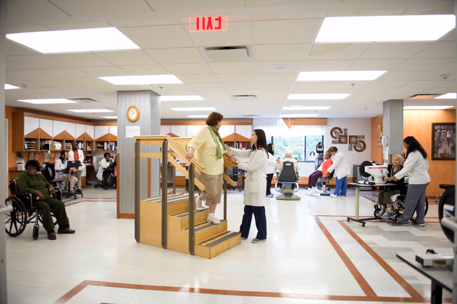 A rehabilitation center room with a healthcare professional assisting a woman walking up a small set of stairs. Other patients and staff are engaged in various physical therapy activities around the room, including seated exercises and wheelchair assistance. The room is well-lit with overhead lights and has a clean, organized appearance with medical equipment and office desks in the background.