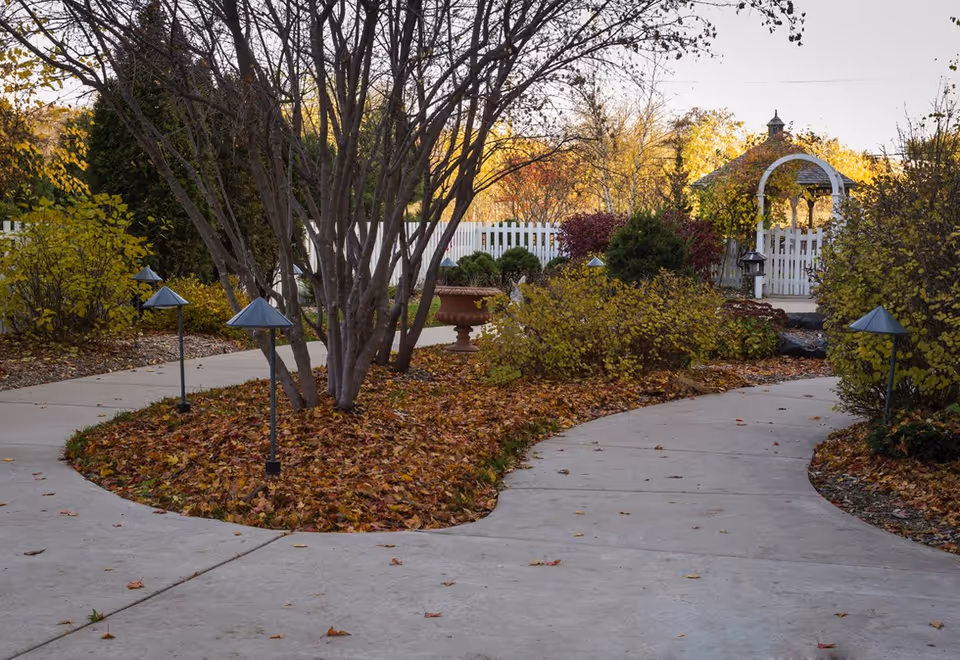Curving concrete walkways through a leaf-strewn garden with small path lights, shrubs, a white picket fence and a gazebo in the background.