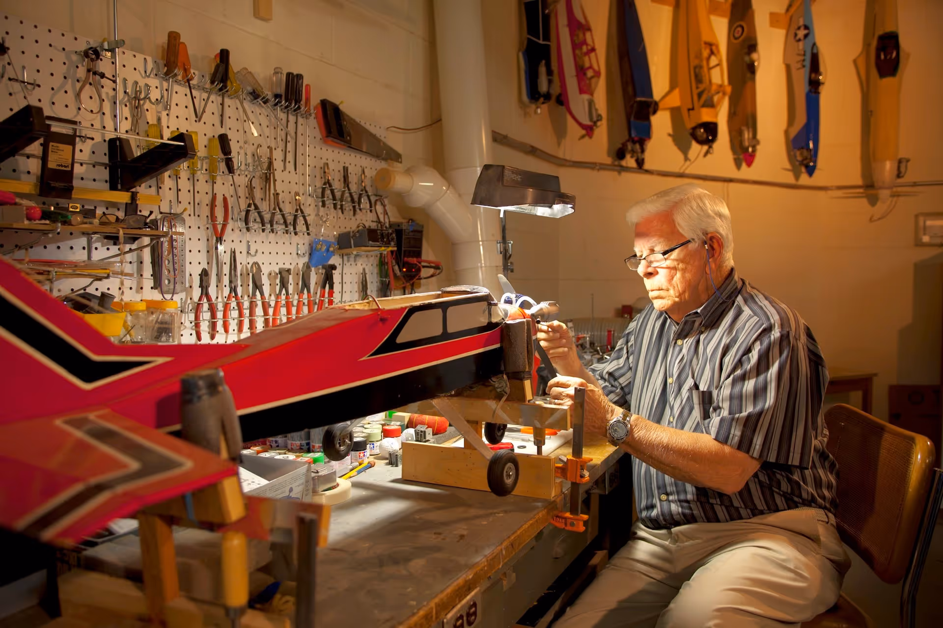 An elderly man wearing glasses and a striped shirt is working on a large red model airplane in a well-lit workshop. The workspace has a pegboard wall with various tools hanging, and several other model airplanes are displayed on the wall behind him.