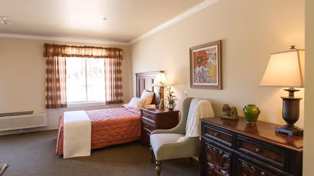 A cozy bedroom in an assisted living facility featuring a bed with a red quilt and white throw, a wooden nightstand with a lamp and plant, a comfortable armchair, a wooden dresser with a lamp and decorative items, a window with plaid curtains, and a framed floral painting on the wall.