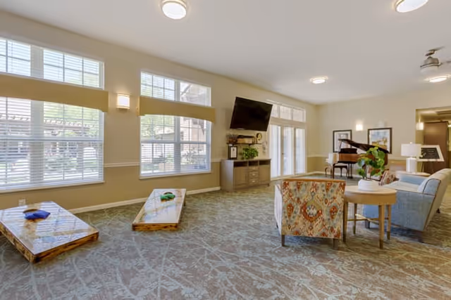 A spacious senior living common area with large windows letting in natural light. The room features a patterned carpet, two wooden cornhole boards with bean bags, a wall-mounted TV above a cabinet with plants, a grand piano in the corner, and comfortable seating including an armchair and a sofa with side tables and lamps.