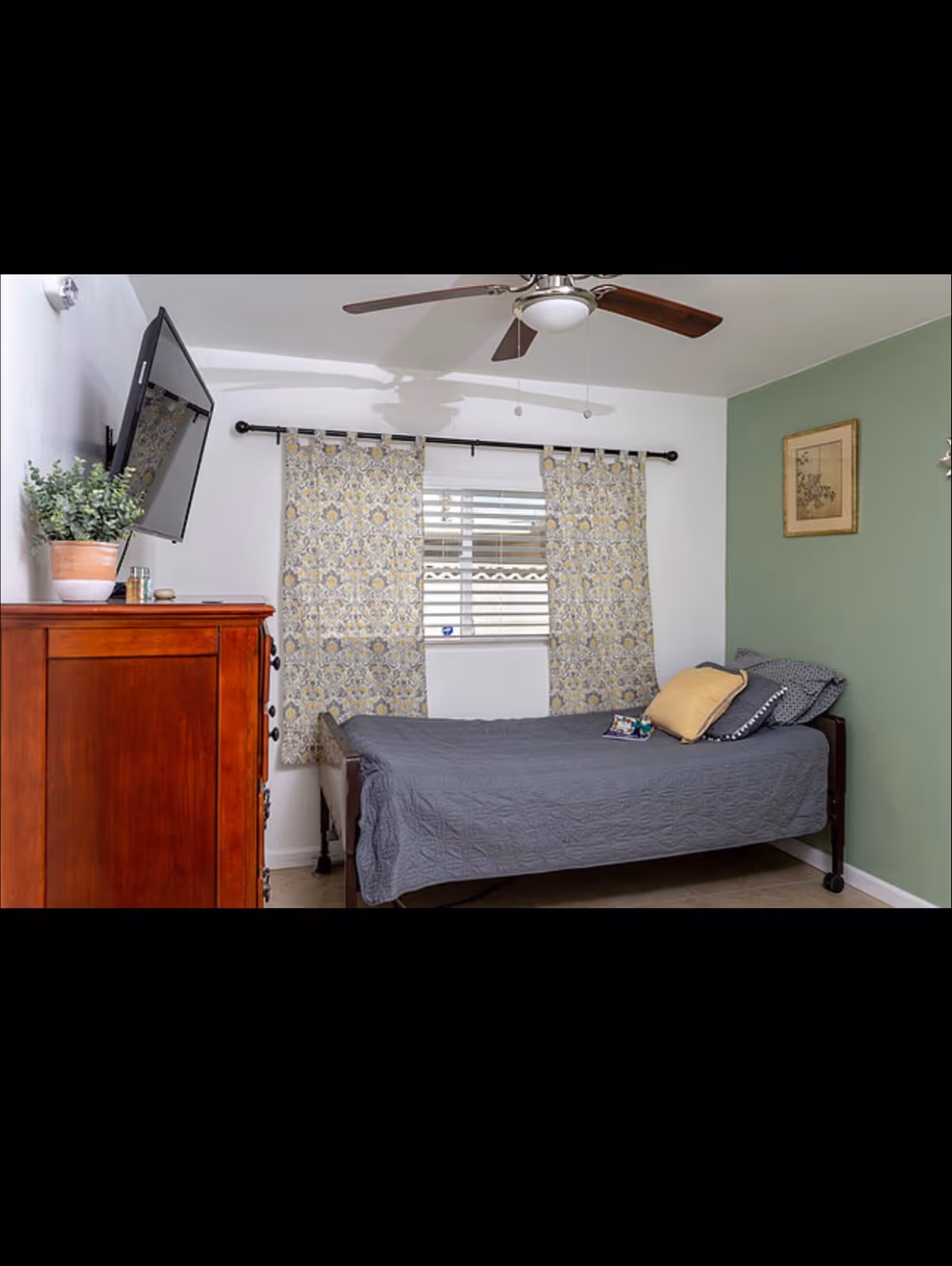 Small bedroom with a single bed, patterned curtains over a window, wall-mounted TV, wooden dresser and a ceiling fan.