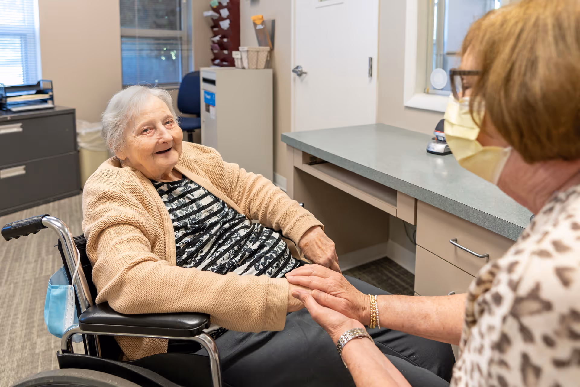 An elderly woman in a wheelchair smiling and holding hands with a caregiver wearing a face mask in a room with office furniture and a window.