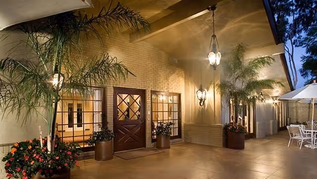 Evening view of an outdoor patio area at Cloisters Of The Valley with potted plants, a wooden door with glass panels, large windows, hanging lantern-style lights, and outdoor seating with a white umbrella.