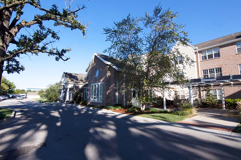 Exterior view of a senior living facility building with brick and siding walls, large windows, a covered patio area with outdoor seating, and trees casting shadows on the driveway under a clear blue sky.