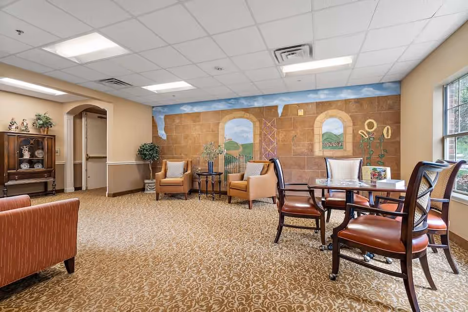 A cozy common area in a senior living facility with patterned carpet, two armchairs with cushions, a small round table with a vase of flowers, and a dining table with four chairs near a large window. The back wall features a mural depicting an outdoor scene with arches, greenery, and sunflowers. There is a wooden cabinet with decorative items on the left side and a doorway leading to another room.