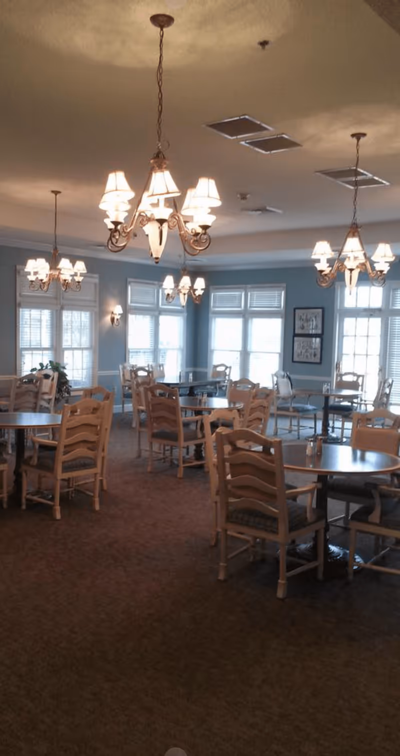 A dining room with multiple round tables and wooden chairs arranged neatly. The room has large windows with white blinds allowing natural light to enter. Several ornate chandeliers with multiple lamps hang from the ceiling, providing warm lighting. The walls are painted a soft blue color, and there are framed pictures on one wall.