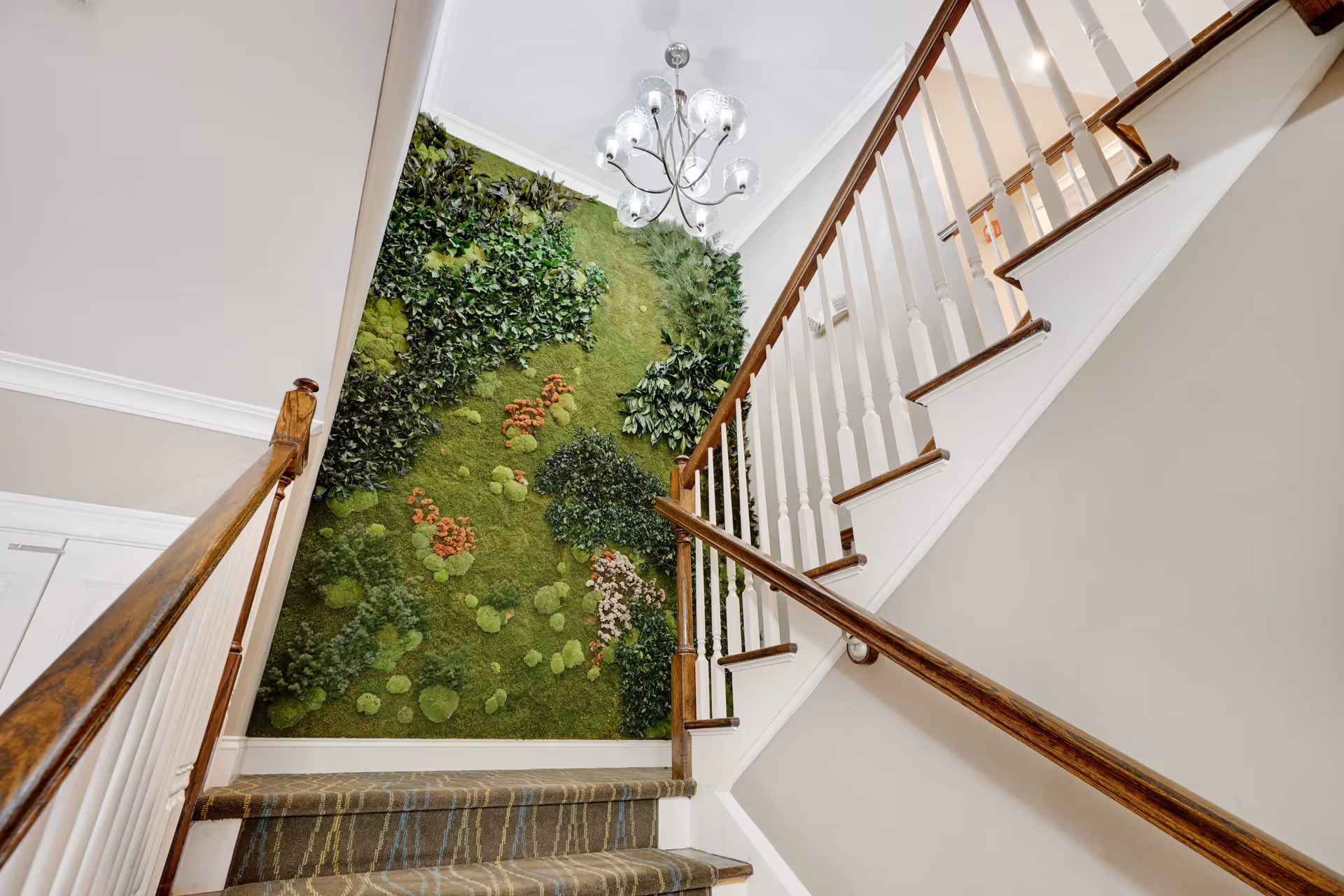 Interior view of a staircase with wooden handrails and white balusters. The wall beside the stairs features a vertical garden with various green plants and moss, accented with some orange and white flowers. A modern chandelier with multiple glass globes hangs from the ceiling above the stairs.