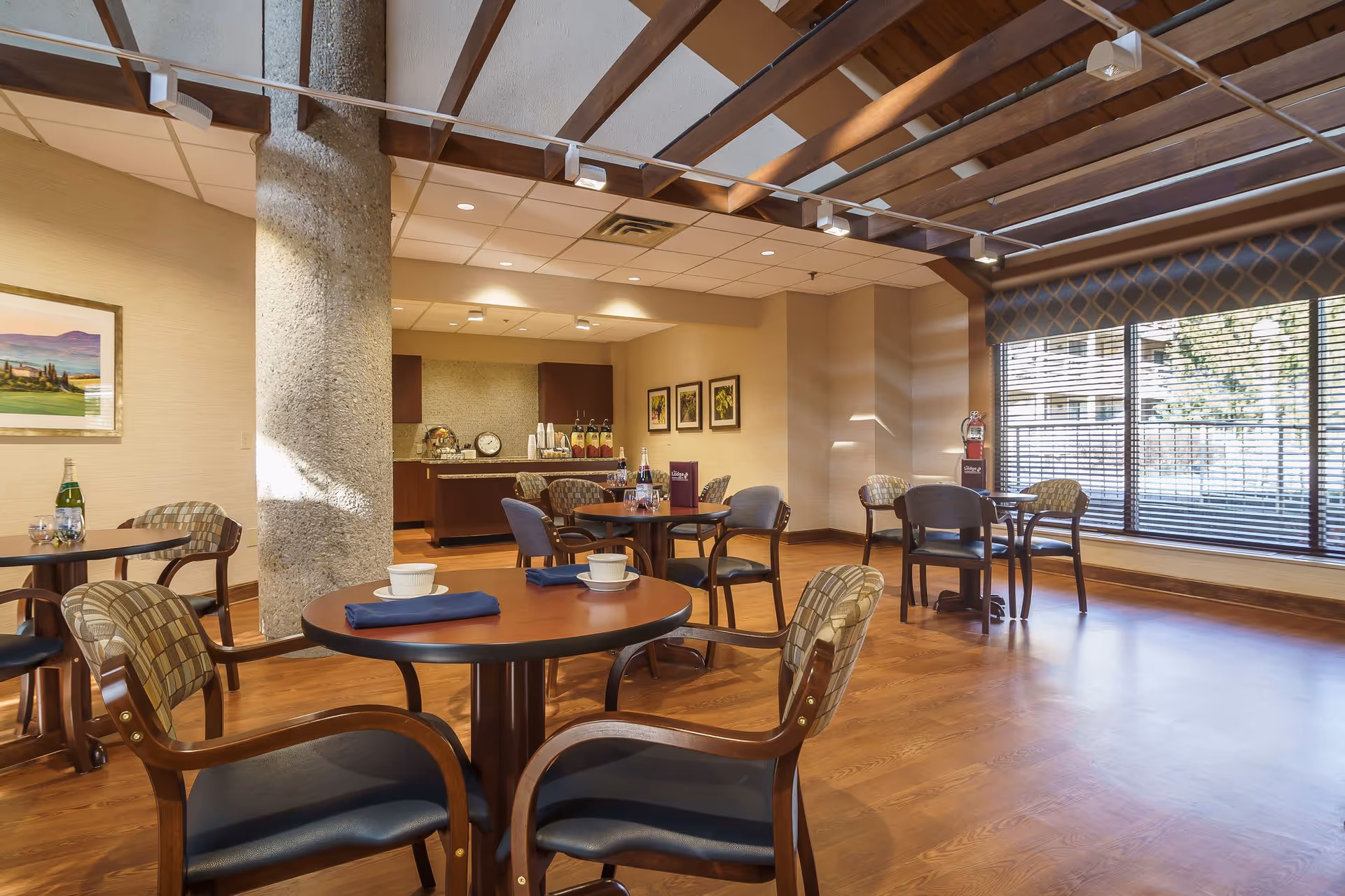 Well-lit communal dining room with round wooden tables and chairs, a beverage station, exposed ceiling beams and large windows.