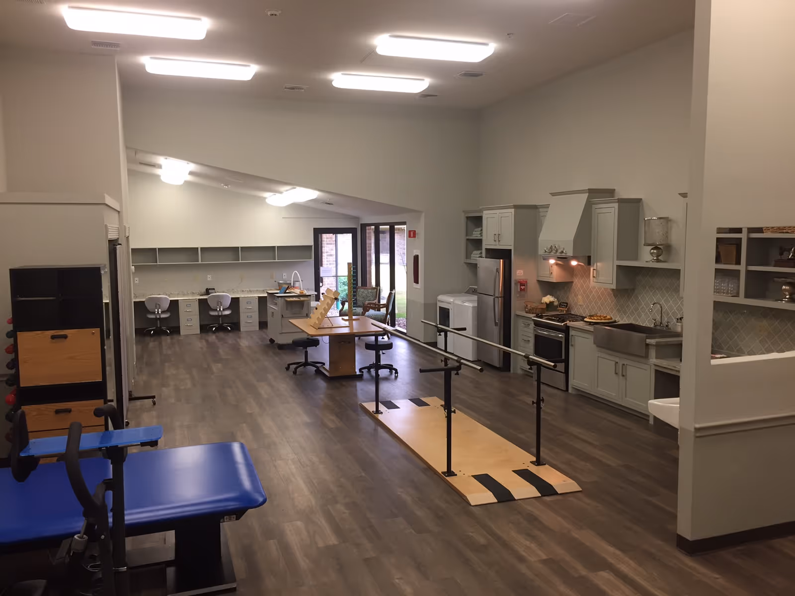 Open interior space with rehabilitation equipment including parallel bars, exercise tables, desks, and a kitchenette.