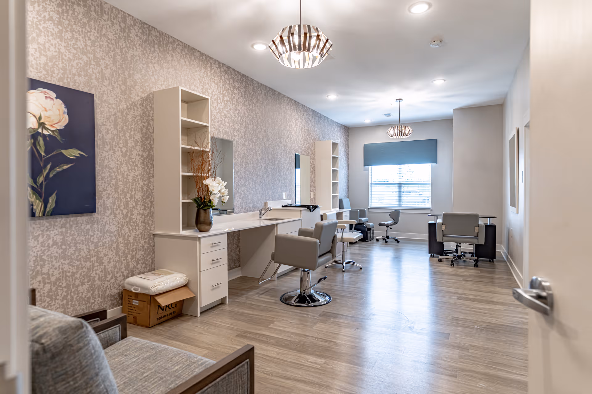 A bright, modern room with light wood flooring and patterned wallpaper. The room features multiple salon-style chairs in front of mirrors and white shelving units. There are two pendant lights hanging from the ceiling, a window with a blue valance, and a desk with an office chair near the window. A floral painting is visible on the left wall, and a cardboard box with folded linens sits on the floor.