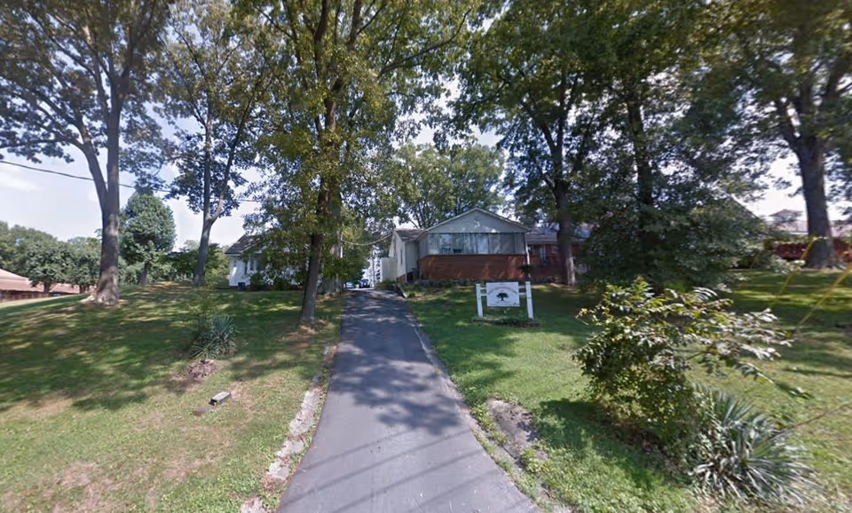 A paved driveway leading uphill to a single-story building surrounded by green grass and tall trees on both sides under a partly cloudy sky.