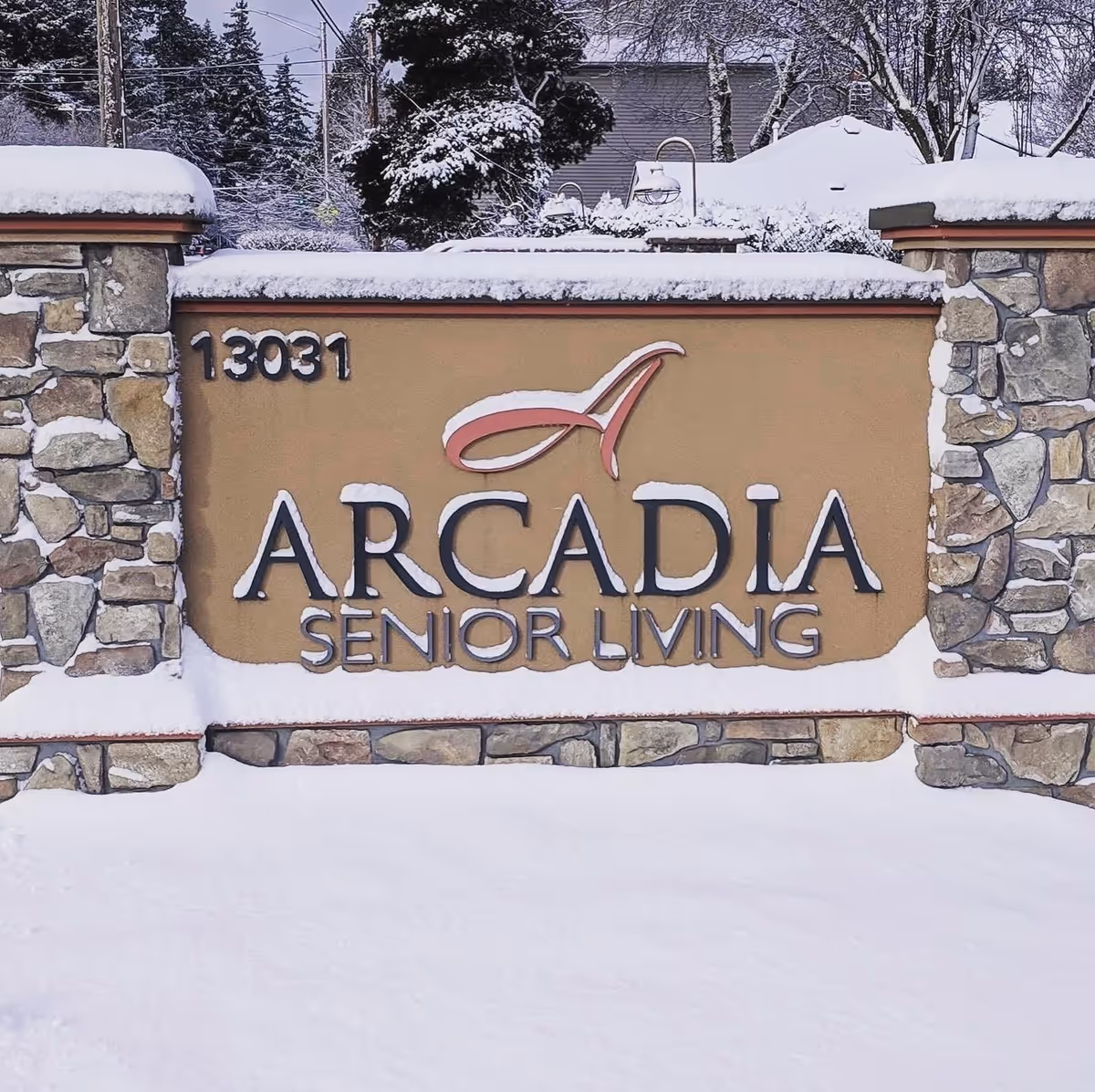 Stone and stucco sign for Arcadia Senior Living covered with snow, with trees and houses in the background also covered in snow.
