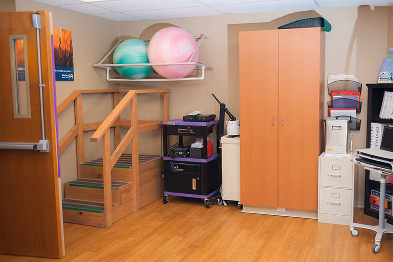 Interior therapy/rehab room with wooden exercise stairs, large therapy balls on a wall rack, a storage cabinet and equipment carts.