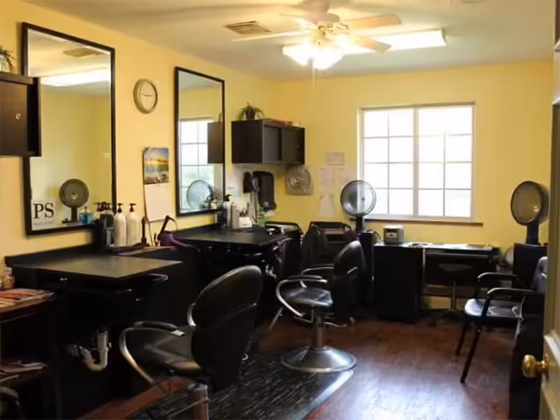 Interior of a salon room with two black salon chairs in front of two large mirrors mounted on a yellow wall. The room has a ceiling fan with lights, a window letting in natural light, and various salon equipment and products on counters and shelves.
