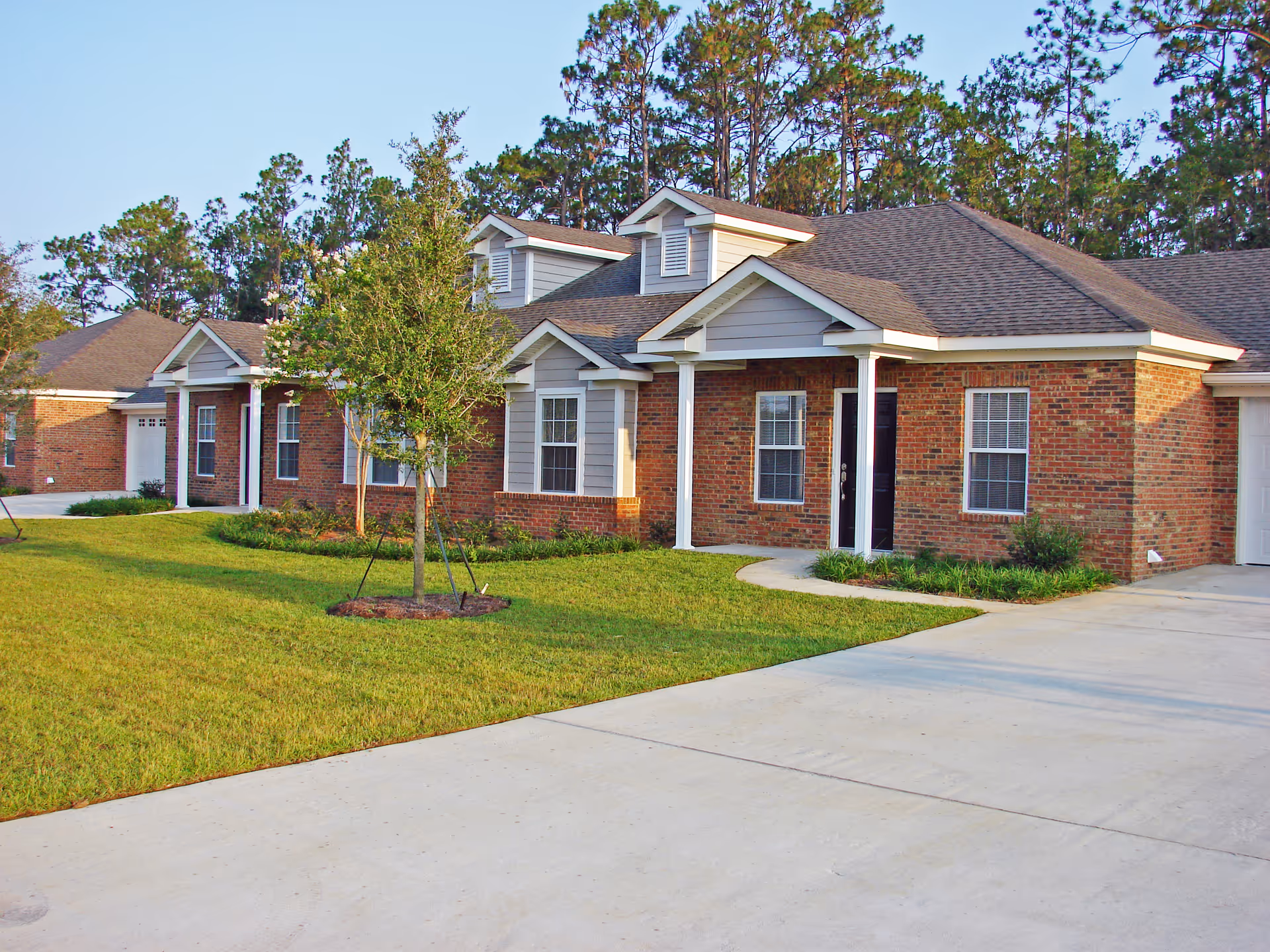 Exterior view of a single-story brick residential building with a well-maintained lawn, small trees, and a concrete driveway. The building has multiple windows and white columns supporting small porch roofs over the entrances.