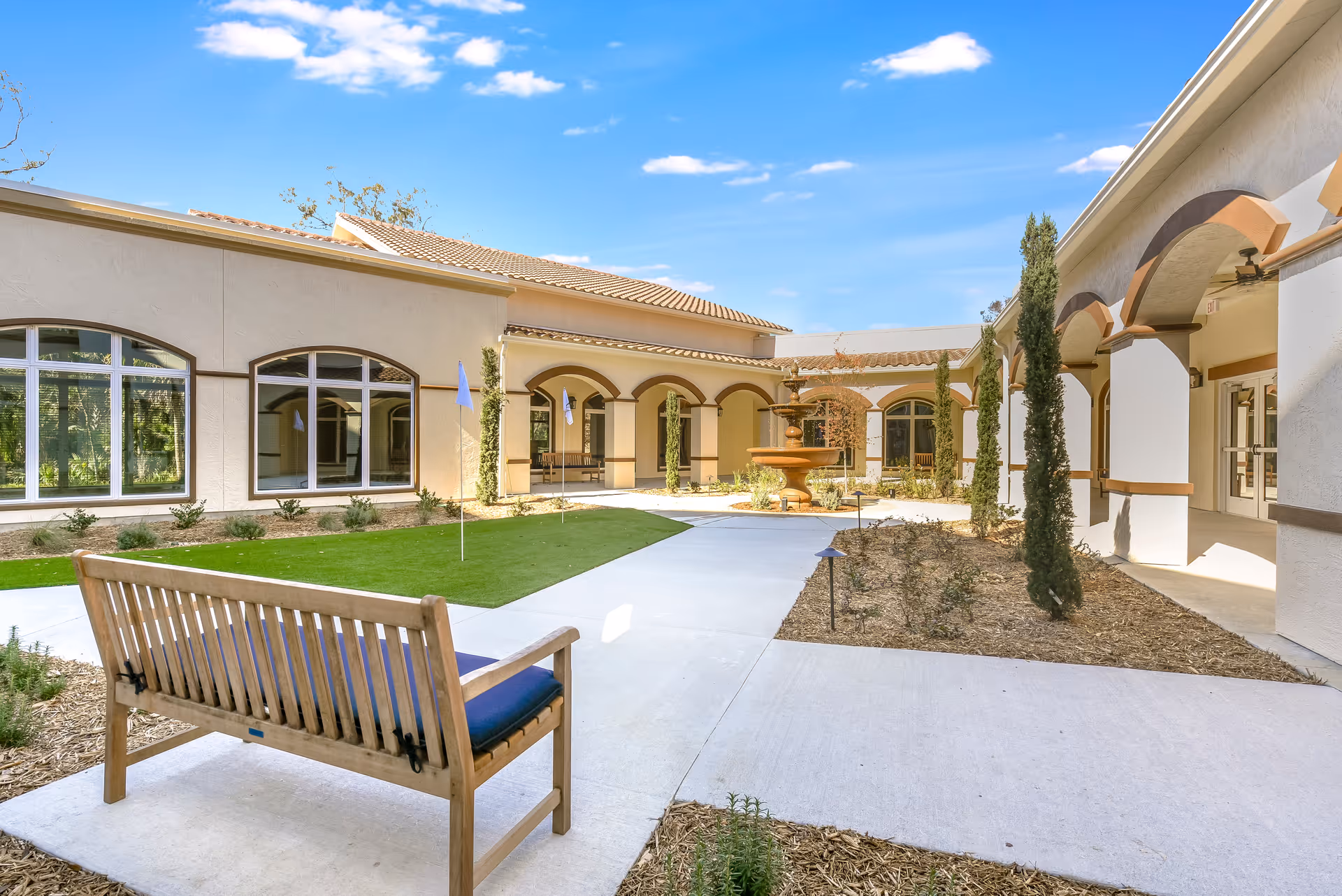 Outdoor courtyard area of Tuscan Gardens of Palm Coast Senior Living featuring a wooden bench with a blue cushion, a putting green with flags, a central fountain, and surrounding beige buildings with arched walkways and tall narrow trees under a blue sky with scattered clouds.