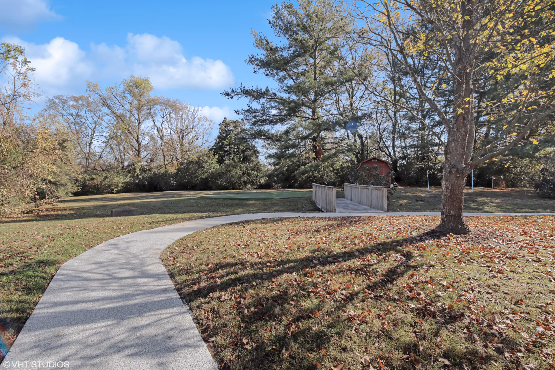 A curved concrete pathway leading through a grassy area with scattered fallen leaves, surrounded by trees with autumn foliage under a partly cloudy blue sky. There is a small wooden bridge and a red shed in the background.