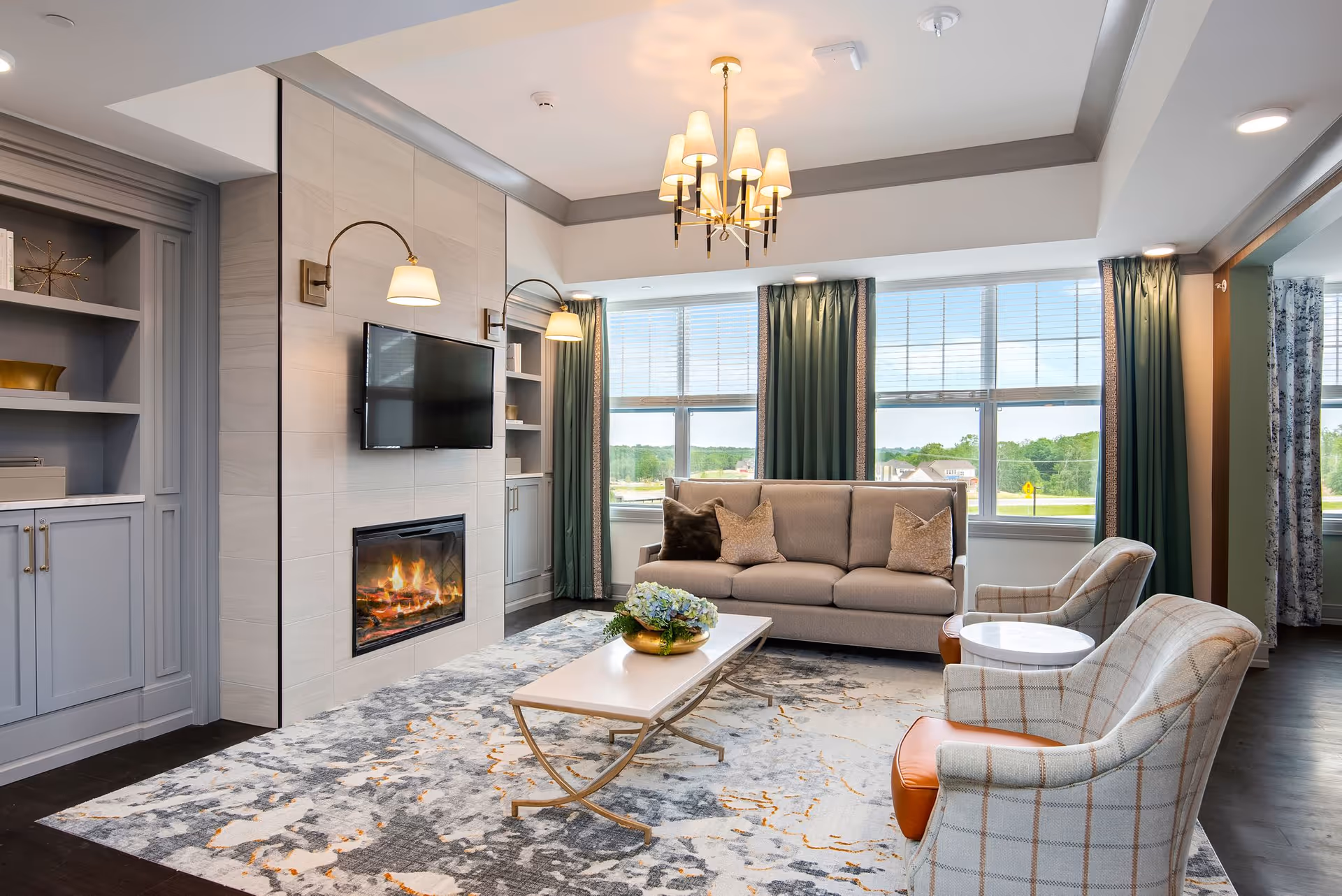 A bright and elegant living room with a beige sofa, two plaid armchairs, a modern fireplace beneath a mounted flat-screen TV, a white coffee table with a floral centerpiece, large windows with green curtains, and built-in shelves and cabinets on the side.