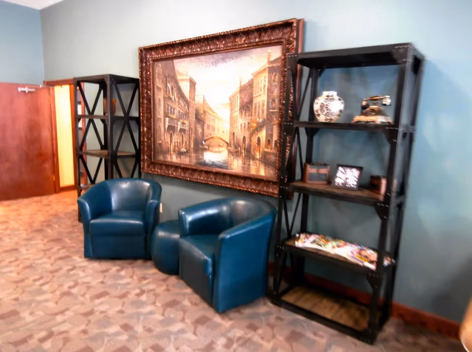 A seating area with two blue leather armchairs and a matching ottoman placed on a patterned carpet. Behind the chairs, there are two black metal shelving units, one of which holds decorative items including a vase, an old-fashioned telephone, and framed pictures. A large framed painting of a European street scene with buildings and a canal hangs on the teal-colored wall.